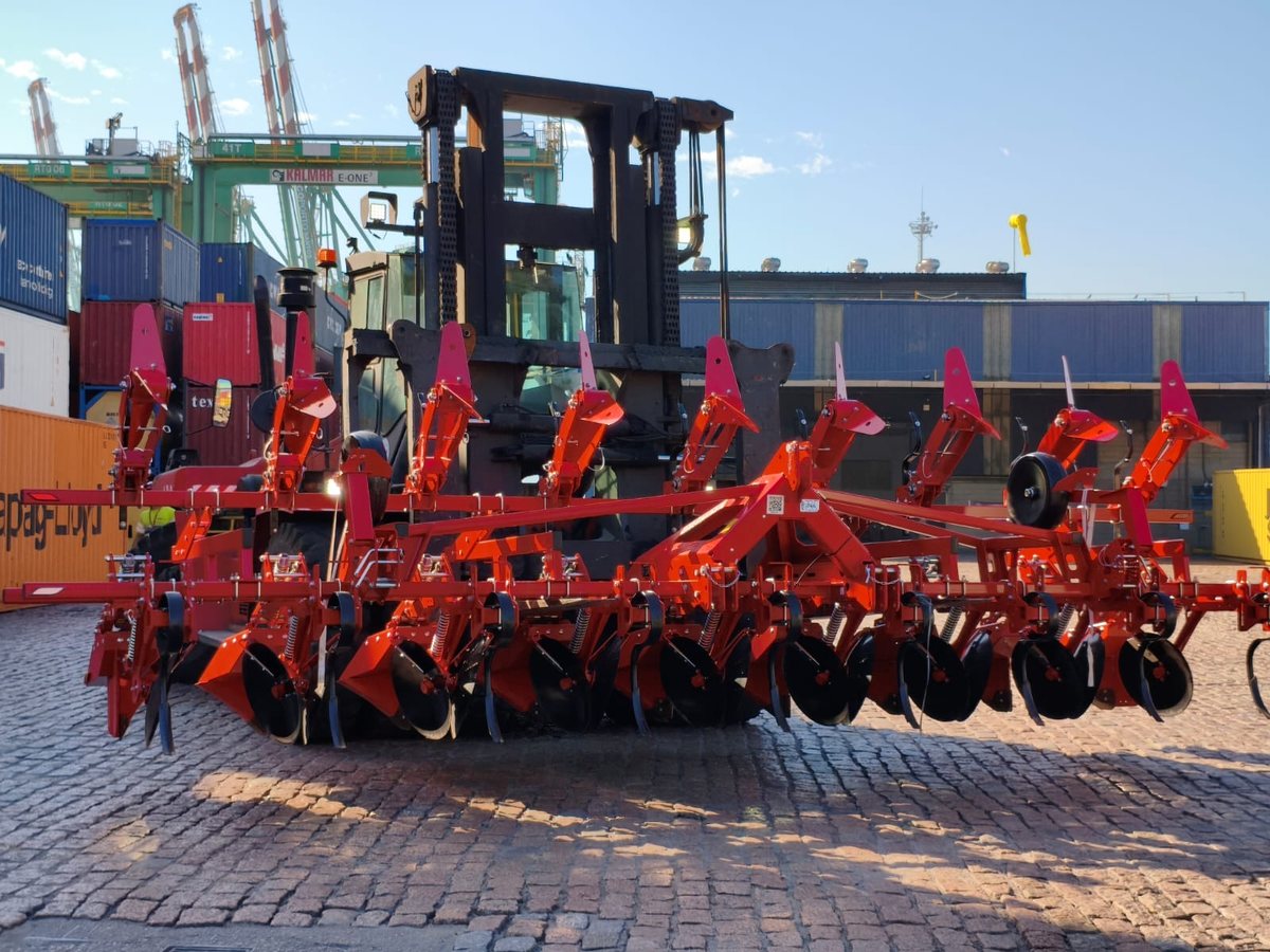 Implemento agrícola vermelho de vários módulos sobre prancha, com contêineres e guindastes ao fundo